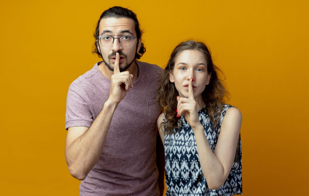 young couple looking at camera making silence gesture with fingers on lips standing over orange background